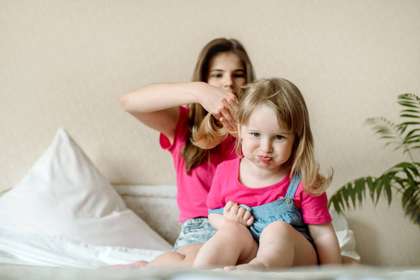 cute sister teen combing hair baby in bed.Cute girls are laughing and smiling. Morning treatments, hair care, care. Family love. Cozy bright bedroom. - 写真・画像