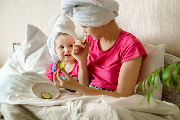 two cute sisters sitting on bed in white towel,applying cucumber slices to her eyes, Baby girls have a funny face.Morning facial, cosmetology. Adult mimicry. Teen first makeup. - Фото, изображение
