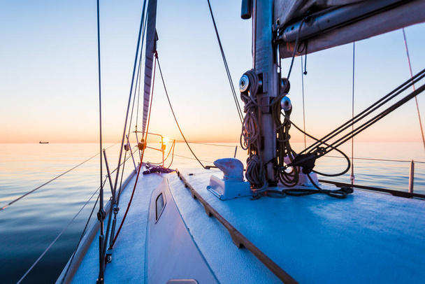 Spokojna woda. Biały jacht żeglujący o zachodzie słońca. Widok z pokładu na dziób, maszt i żagle. Morze Bałtyckie, Łotwa - Zdjęcie, obraz