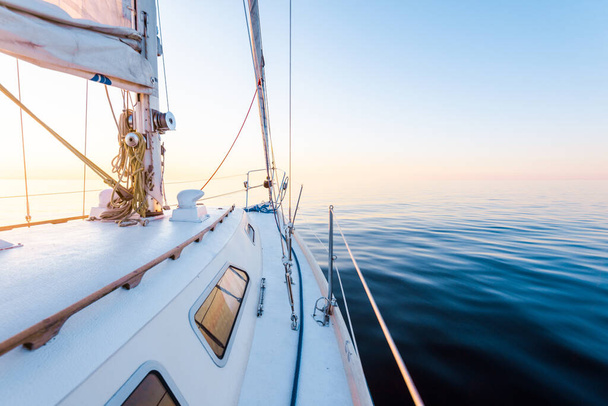 Spokojna woda. Biały sloop sfałszował jacht o zachodzie słońca. Widok z pokładu na dziób, maszt i żagle. Morze Bałtyckie, Łotwa - Zdjęcie, obraz