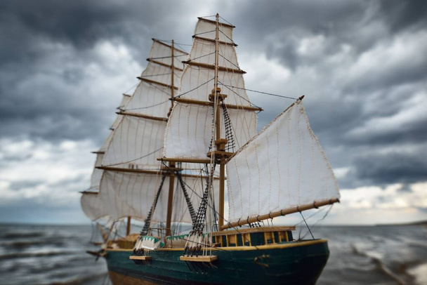 Antiquarian wooden scale model of the clipper tall ship, close-up. Dramatic sky, dark stormy clouds and sea. Traditional craft, souvenir, hobby, collecting, vintage, modeling. Zero waste concept - Photo, Image