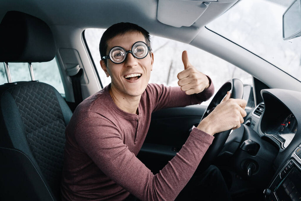Drôle et ridicule idiot nerd driver dans de grandes lunettes tenant le volant et souriant à la caméra. Concept d'un conducteur débutant distrait de la route - Photo, image