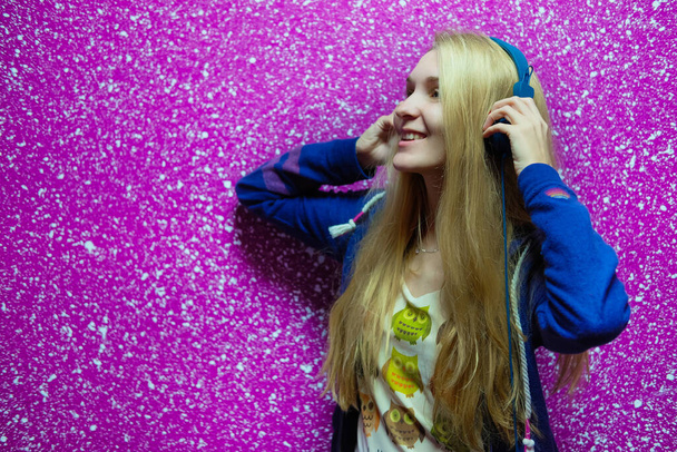 chica rubia en una sudadera con capucha púrpura escuchando música en auriculares azules sobre fondo rosa. hipster adolescente disfruta escuchando el audio libro con los ojos cerrados. sudaderas - Foto, Imagen