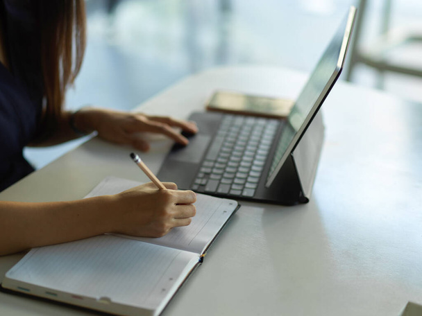 Vista lateral del trabajador de oficina femenino torcer a mano en el cuaderno mientras trabaja con la tableta digital  - Foto, Imagen
