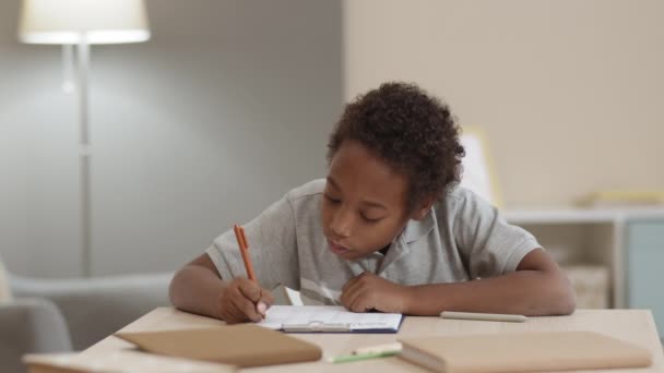 自宅の机の上に座って紙の上にペンで書いて考えながら10歳のアフリカの男性の子供の中程度のクローズアップ。宿題をする生徒 - 映像、動画