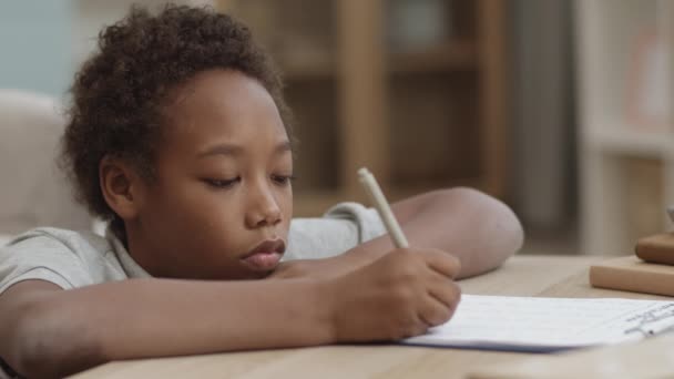 Gros plan d'un garçon africain de dix ans qui fait ses devoirs dans une pièce par bureau, écrit avec un stylo sur une feuille de papier et se gratte la tête - Séquence, vidéo