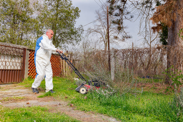 Ogrodnik noszący ochronne ogólnie jest cięcie trawy w jego podwórku z kosiarką silnikową wśród drzew owocowych. - Zdjęcie, obraz