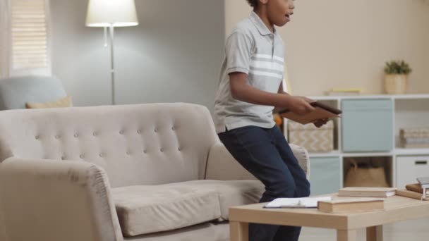 Μεσαίο μήκος της Αφρικής σχολείο αγόρι τρέχει στον καναπέ, κάθεται σε αυτό, χρησιμοποιώντας το tablet υπολογιστή. Παιδί απολαμβάνει ελεύθερο χρόνο στο σαλόνι στο σπίτι - Πλάνα, βίντεο