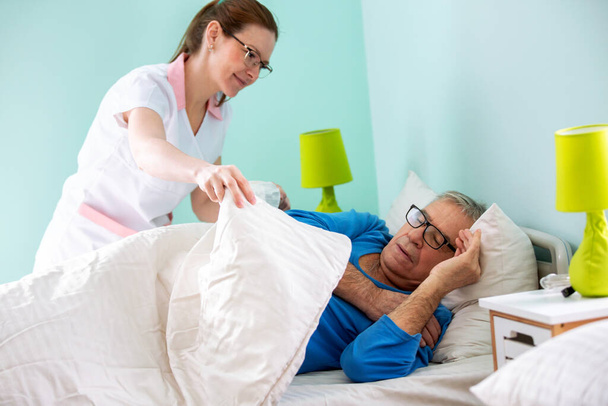 Elder man resident of a nursing home sleeping while medical staff nurse is covering him with sheet, elder people care concept - Photo, Image
