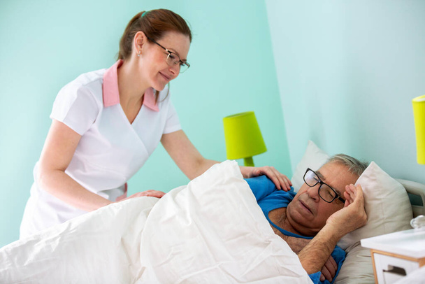 Elder man resident of a nursing home sleeping while medical staff nurse is covering him with sheet, elder people care concept - Photo, Image