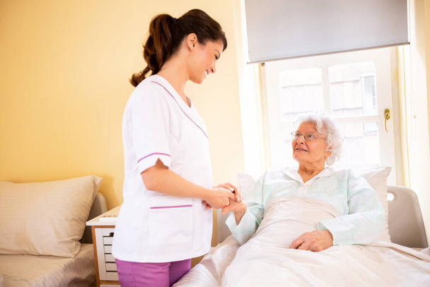 Nursing home doctor holding a hand of a senior woman patient occupant while she is lying on the bed - Foto, Imagem