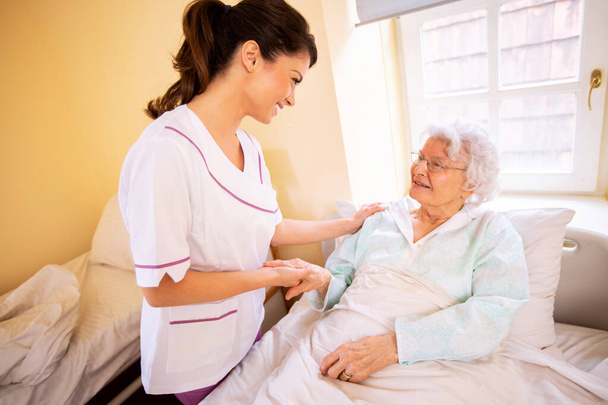Nursing home doctor holding a hand of a senior woman patient occupant while she is lying on the bed - Foto, Imagen