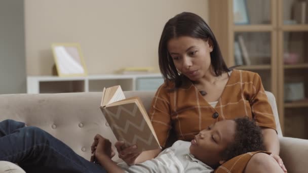 Mittlerer afrikanischer Schuljunge liegt im Wohnzimmer auf dem Sofa auf Knien einer jungen Mutter mit gemischter Rasse, liest ihm laut vor, lächelt - Filmmaterial, Video