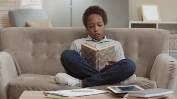 Volle Aufnahme eines fröhlichen afrikanischen Schuljungen in Jeans und Poloshirt, der in Lotus-Pose auf dem Sofa im Wohnzimmer sitzt und sich laut Buch vorliest - Filmmaterial, Video