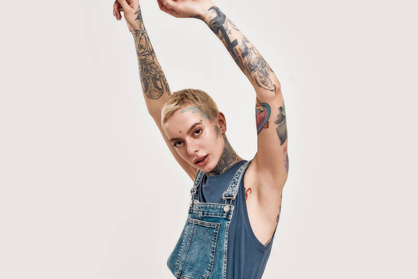 Tattoo and piercing. A white tattooed and pierced woman dancing alone with her hands raised up wearing a denim overall looking into a camera - Fotografie, Obrázek