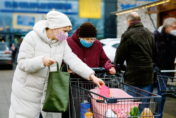 Senior woman and social worker with medical mask due pandemic coronavirus disease. Daughter or granddaughter help grandmother with shopping in supermarket, push cart trolley with foods, outdoors - Photo, Image
