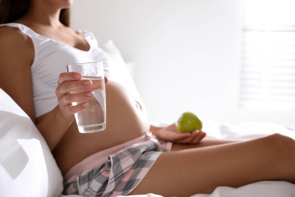 Young pregnant woman with glass of water and apple in bedroom, closeup. Taking care of baby health - Fotó, kép
