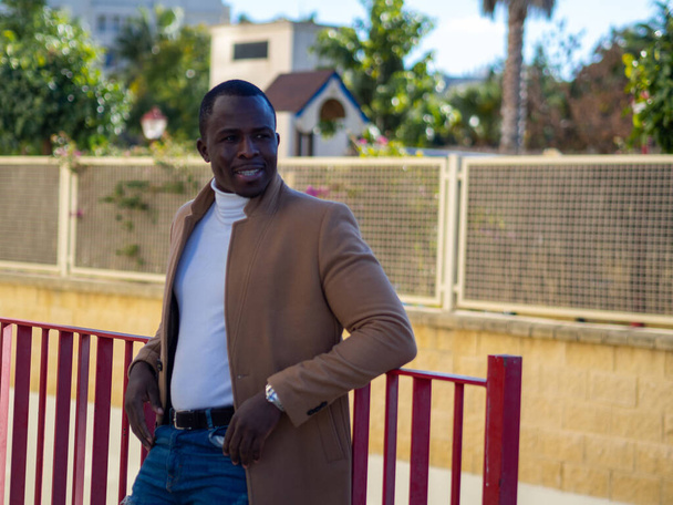 A shallow focus of a stylish African male wearing jeans with a white sweater and beige coat and leaning on a fence - Foto, Imagem