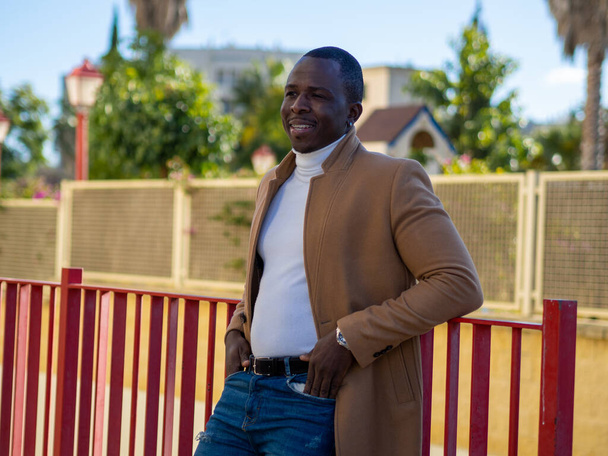 A shallow focus of a stylish African male wearing jeans with a white sweater and beige coat and leaning on a fence - Foto, Bild