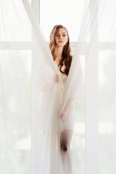 woman in white robe stands on windowsill. morning of bride. reflection in glass - Φωτογραφία, εικόνα