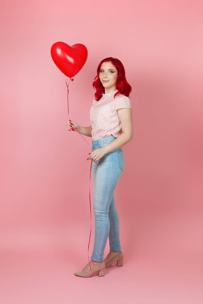 full-length happy, slender young woman with red hair in jeans holds a large flying red balloon in the shape of a heart, isolated on a pink background - Fotó, kép