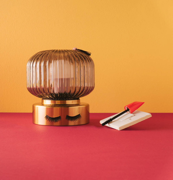 Lamp with the fake eyelashes and mascara on light switch. Peachy yellow background. - Photo, Image