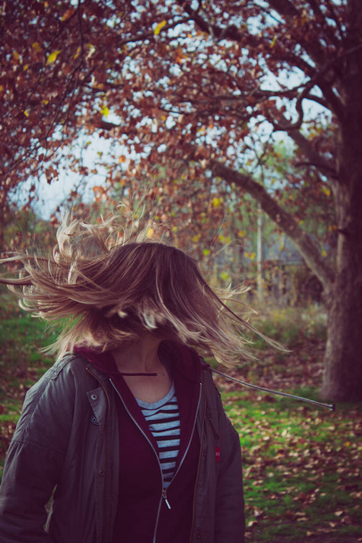 A young woman looking to her back while her hair flips with an autumn tree in the background - Photo, Image
