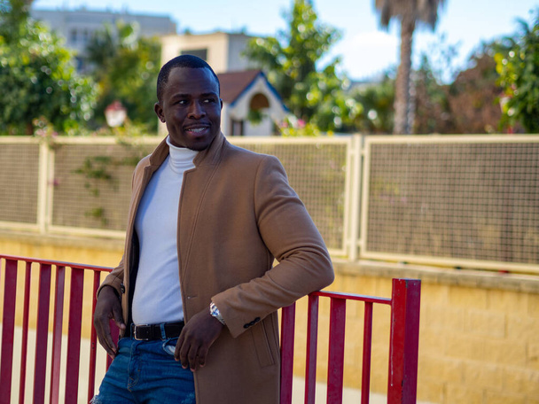 A shallow focus of a stylish African male wearing jeans with a white sweater and beige coat and leaning on a fence - Фото, изображение