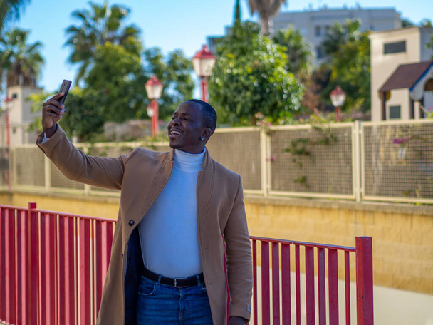 A shallow focus of a cheerful African male video calling with a phone outdoors - 写真・画像