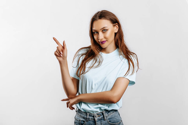 Portrait of a smiling girl pointing finger to the side at on a white isolated background. A woman points to an idea, a place for advertising. Positive girl with fashion make-up with purple lips. - Foto, afbeelding