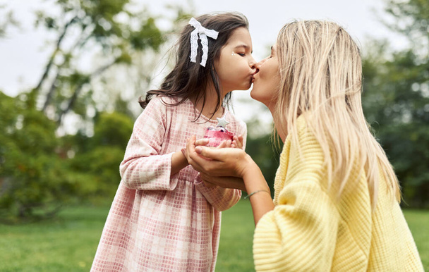 A happy daughter celebrates her birthday with her family in the park. A beautiful young woman brought a birthday cake with a candle for her little girl outdoor. Happy Mother's day and birthday concept - Foto, Imagem