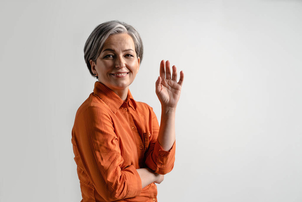 Middle aged grey haired pretty woman smile gentle looking at camera with touching finger tips wearing orange shirt and denim jeans isolated on white background. Human emotions concept - Foto, immagini