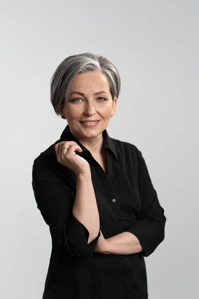 Grey haired mid aged pretty woman looking at camera with arms folded wearing black shirt isolated on grey background. Confident mature grey haired woman. Human emotions, facial expression concept - Foto, imagen