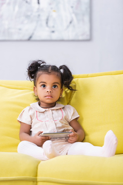 toddler african american girl holding smartphone while sitting on sofa - Foto, imagen