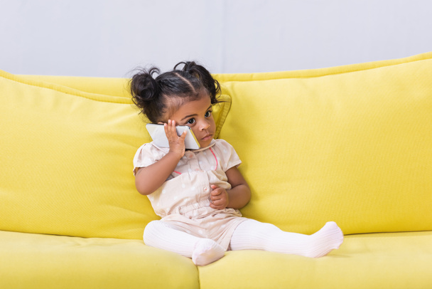 Africana niña americana hablando en el teléfono inteligente mientras está sentado en el sofá - Foto, Imagen