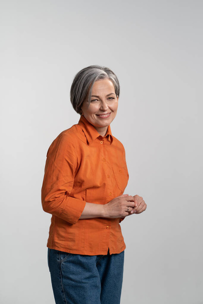 Middle aged grey haired pretty woman smile gentle looking at camera with touching finger tips wearing orange shirt and denim jeans isolated on white background. Human emotions concept - Valokuva, kuva