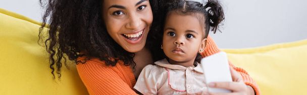 счастливая африканская американская мать держит смартфон рядом с ребенком малыша, баннер - Фото, изображение
