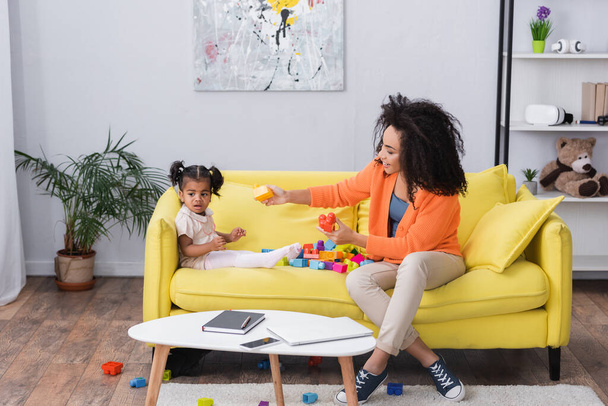 happy african american mother playing building blocks with toddler kid on sofa - Foto, afbeelding