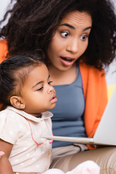 amazed african american freelancer using laptop near toddler daughter in living room - Foto, Bild