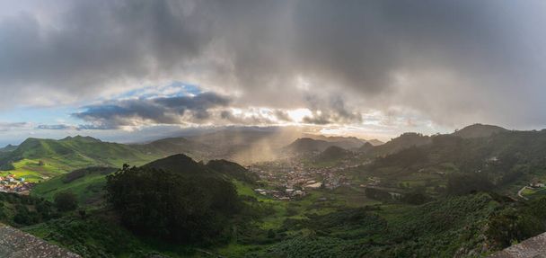 Village vu d'un point de vue au milieu de montagnes verdoyantes avec une journée nuageuse et des rayons du soleil - Photo, image