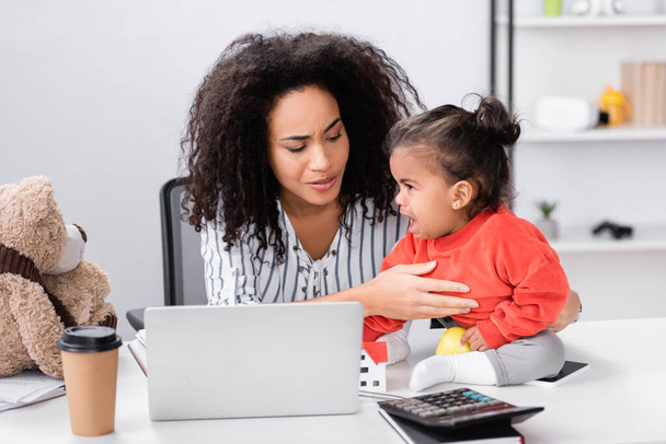 curly african american freelancer supporting toddler daughter sitting on desk near laptop  - Zdjęcie, obraz