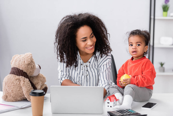 happy african american mother looking at toddler daughter sitting on desk with apple near gadgets - Фото, изображение