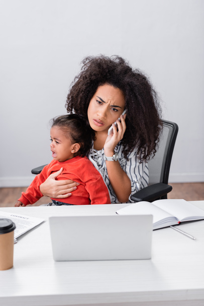 stresszes afro-amerikai szabadúszó gazdaság karokban sírás kisgyermek lánya, miközben beszél okostelefonon  - Fotó, kép