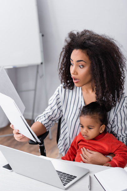surprised african american mother holding folder while sitting with toddler daughter and working from home  - Foto, immagini