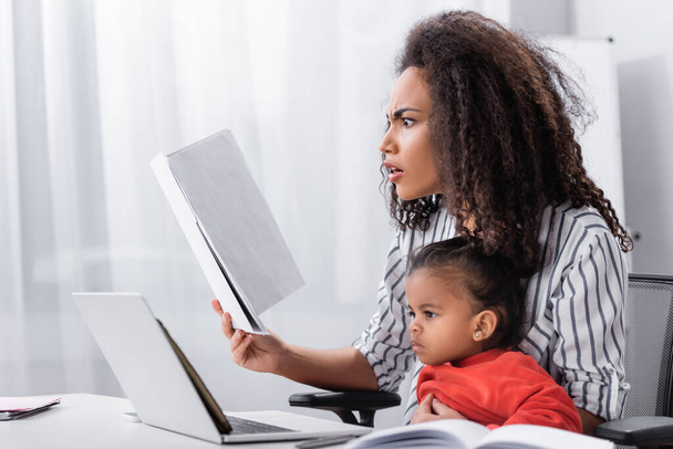 stressed african american mother holding folder while sitting with toddler daughter and working from home  - Фото, изображение