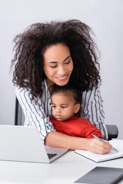 happy african american mother sitting with toddler daughter while writing in notebook neat laptop on desk - 写真・画像