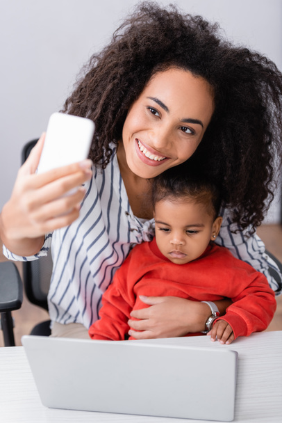 happy african american mother taking selfie with toddler daughter near laptop on blurred foreground - Photo, Image