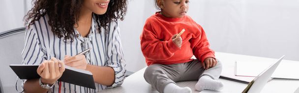 peuter afrikaans amerikaans kind houden smakelijk koekje en zitten op bureau in de buurt van moeder schrijven in notebook in de buurt van laptop, banner - Foto, afbeelding