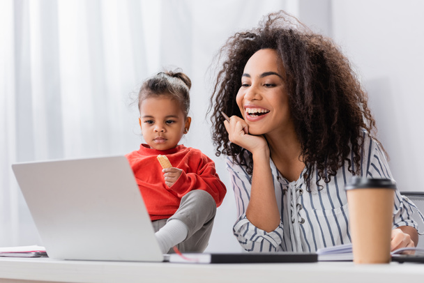 toddler african american kid holding tasty cookie and sitting on desk near happy mother looking at laptop - Photo, Image