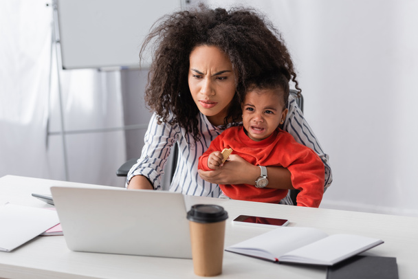 estresada madre afroamericana sosteniendo en brazos llorando niño pequeño mientras trabaja desde casa  - Foto, imagen